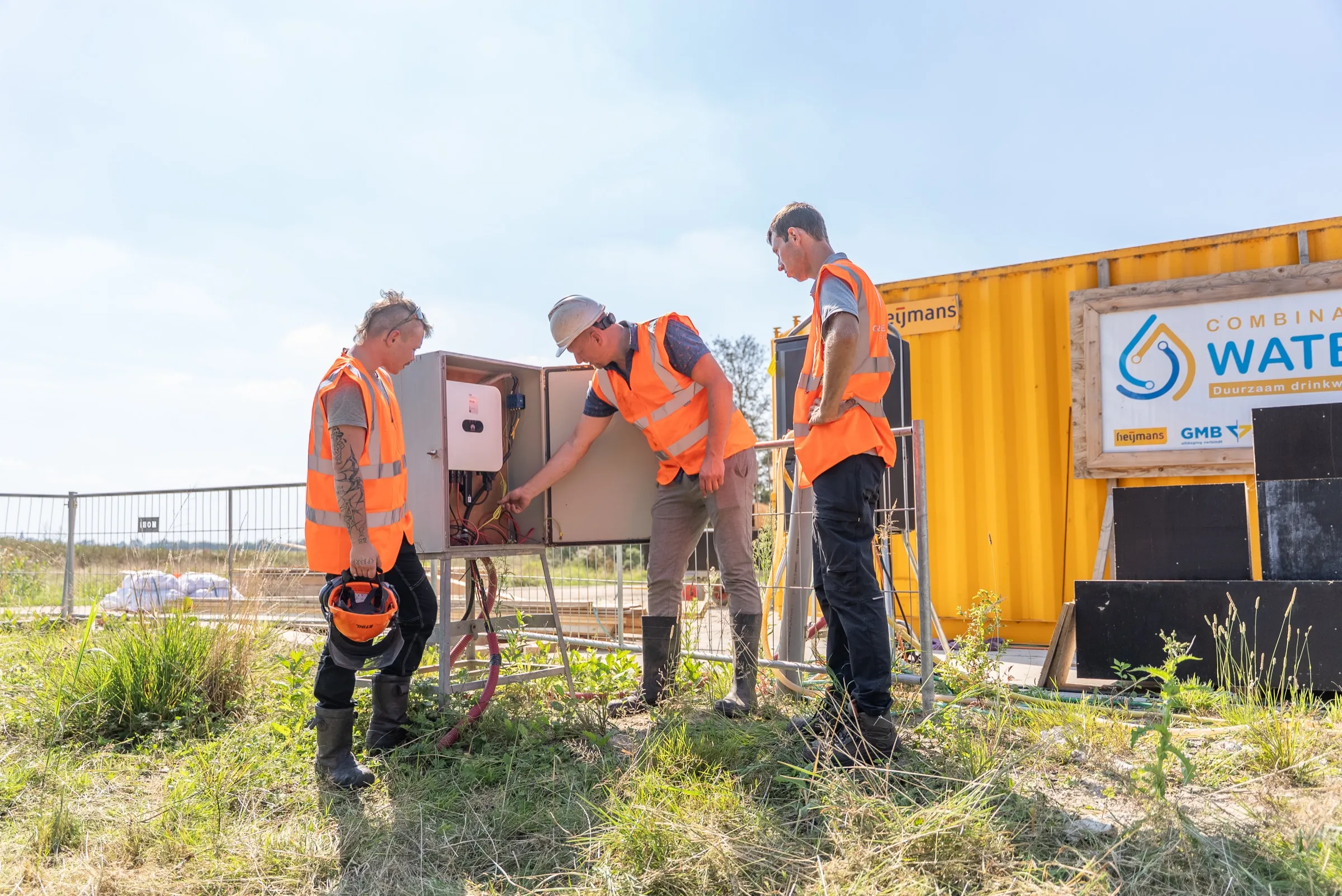 Drie medewerkers bespreken de omvormerkast naast een gele container op een bouwterrein